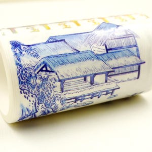 Puede incluir: Un rollo de cinta washi azul y blanca con un diseño de una casa japonesa con techo de paja y un puente. La cinta está sobre un fondo blanco.