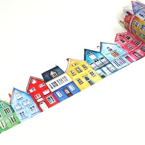 May include: A roll of decorative washi tape featuring a colorful illustration of quaint houses. The houses are painted in various colors, including green, blue, red, and yellow, with detailed windows and architectural features. The tape is unrolled, showing the repeating pattern.