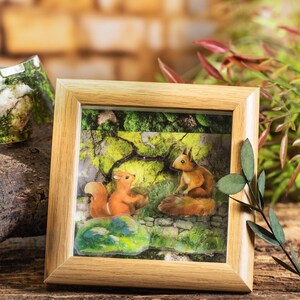 Peut inclure: Une sc&egrave;ne de for&ecirc;t encadr&eacute;e pr&eacute;sente deux &eacute;cureuils roux avec des queues touffues. L'un tient un gland, l'autre est assis sur un rocher. L'arri&egrave;re-plan repr&eacute;sente une for&ecirc;t avec un feuillage vert et un mur en pierre. Le cadre est en bois clair.