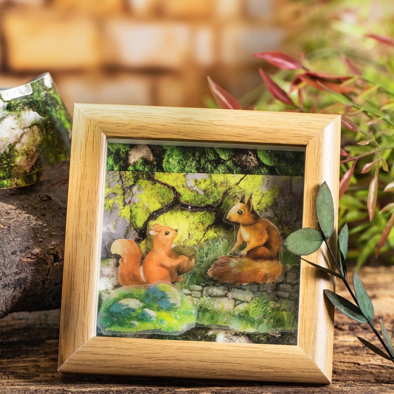 Peut inclure: Une sc&egrave;ne de for&ecirc;t encadr&eacute;e pr&eacute;sente deux &eacute;cureuils roux avec des queues touffues. L'un tient un gland, l'autre est assis sur un rocher. L'arri&egrave;re-plan repr&eacute;sente une for&ecirc;t avec un feuillage vert et un mur en pierre. Le cadre est en bois clair.