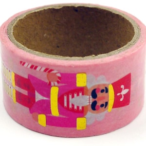 Peut inclure: Un rouleau de ruban adhésif décoratif rose orné d'un motif répétitif de soldat casse-noisette. Le casse-noisette est illustré dans des tons de rose, rouge et jaune, portant un haut chapeau avec une fleur de lys blanche et tenant une sucette.