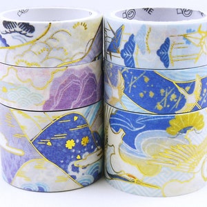 May include: Two stacks of decorative washi tape rolls. The tape features Japanese-inspired designs with gold accents, including cranes, mountains, and floral patterns. The color palette includes blue, purple, and white.