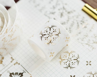 May include: A roll of white washi tape with gold floral and geometric patterns. The tape is partially unrolled, revealing more of the patterned design. The background features similar designs on a grid.