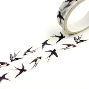Könnte beinhalten: Dekoratives Washi-Tape mit einem sich wiederholenden Muster aus schwarzen und weißen Schwalben im Flug. Die Tape-Rolle ist weiß mit einem grau-schwarzen Vogeldesign. Das Tape ist abgerollt und zeigt das Vogelmuster.
