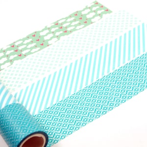 Puede incluir: Cuatro rollos de cinta adhesiva decorativa dispuestos en fila. Los patrones incluyen nubes blancas con pequeños corazones rojos sobre fondo verde claro, una cuadrícula de rombos blancos sobre menta, rayas diagonales turquesas y un patrón geométrico azul cerceta.