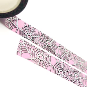 May include: Pink and black patterned washi tape with a spiderweb design. The tape is rolled up and partially unrolled, showing the pattern.