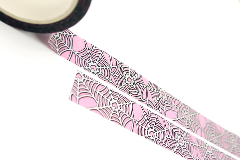 May include: Pink and black patterned washi tape with a spiderweb design. The tape is rolled up and partially unrolled, showing the pattern.