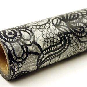 Puede incluir: Un rollo de cinta washi decorativa con un diseño de encaje negro y gris. La cinta está enrollada, mostrando el intrincado patrón de encaje sobre un fondo gris claro. Esta cinta de papel se utiliza para manualidades y decoración.