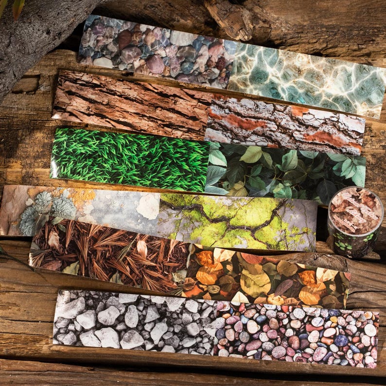 Peut inclure: Une collection de rubans adh&eacute;sifs d&eacute;coratifs washi avec des motifs inspir&eacute;s de la nature, notamment de l'&eacute;corce, de l'herbe, des feuilles, des pierres et de l'eau. Les rubans sont dispos&eacute;s sur une surface en bois, mettant en valeur leurs textures et couleurs vari&eacute;es.