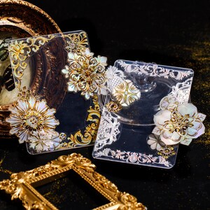 May include: Two clear acrylic trays with ornate gold and silver designs. The trays feature decorative, iridescent, shell-like flowers with gold accents.