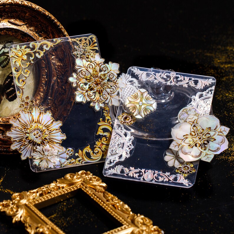 May include: Two clear acrylic trays with ornate gold and silver designs. The trays feature decorative, iridescent, shell-like flowers with gold accents.