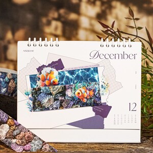 Peut inclure: Calendrier de bureau blanc pour d&eacute;cembre, reli&eacute; par spirale, avec des images sous-marines de poissons-clowns et de roches. Le calendrier a un design violet et le num&eacute;ro 12 est visible.
