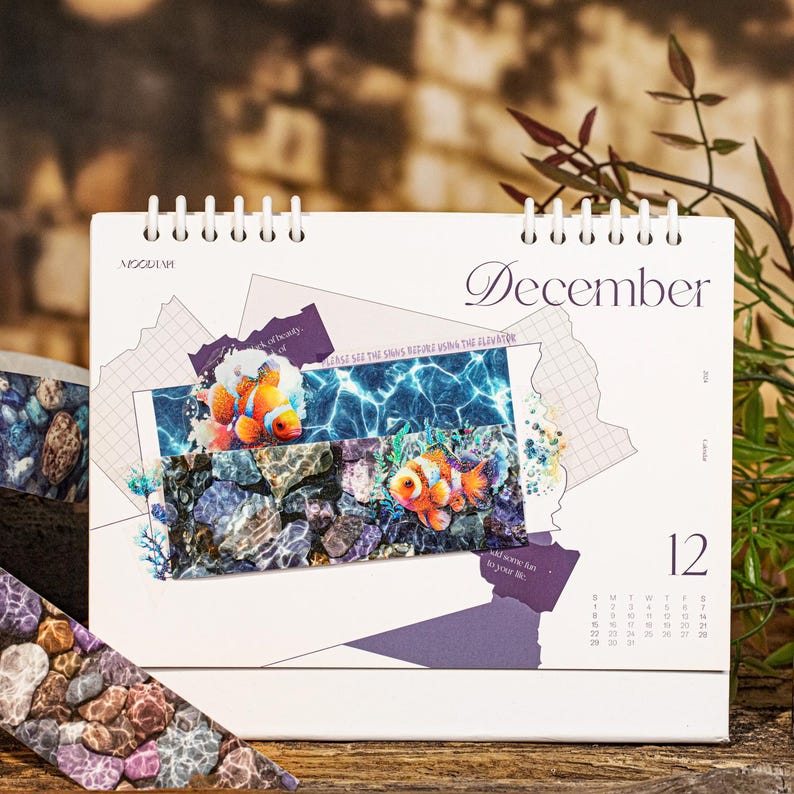 Peut inclure: Calendrier de bureau blanc pour d&eacute;cembre, reli&eacute; par spirale, avec des images sous-marines de poissons-clowns et de roches. Le calendrier a un design violet et le num&eacute;ro 12 est visible.