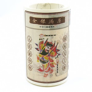 Puede incluir: Un recipiente cilíndrico con una etiqueta beige que presenta caracteres chinos y una colorida ilustración de una deidad. La etiqueta incluye texto en chino y un borde decorativo. El recipiente es blanco.