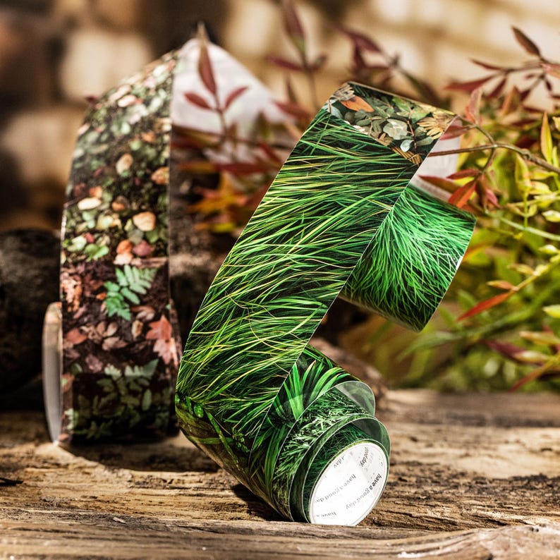 Peut inclure: Trois rouleaux de ruban adh&eacute;sif d&eacute;coratif washi avec des motifs sur le th&egrave;me de la nature. Un rouleau pr&eacute;sente un motif d'herbe verte, un autre avec un motif de feuilles et de feuillage, et un troisi&egrave;me avec un motif forestier similaire. Le ruban est pr&eacute;sent&eacute; sur une surface en bois.