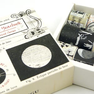May include: A white box set containing rolls of decorative washi tape with black and white celestial designs. The box next to it is labelled "Astrological Yearbook" with diagrams of the moon phases.