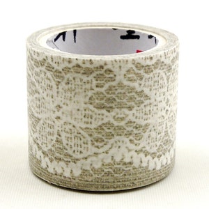 Può includere: Un rotolo di nastro adesivo decorativo con un motivo a pizzo bianco e beige. Il nastro è cilindrico e poggia su una superficie bianca. Il nucleo interno del rotolo di nastro è visibile.
