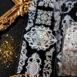 May include: A roll of decorative adhesive tape with a silver iridescent finish. The tape features a repeating pattern of ornate, intricate designs in a floral and geometric style.