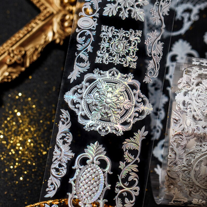 May include: A roll of decorative adhesive tape with a silver iridescent finish. The tape features a repeating pattern of ornate, intricate designs in a floral and geometric style.