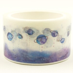 Op de afbeelding: Een rol decoratieve plakband met een ruimte-thema in aquarel-stijl. Het ontwerp toont diverse blauwe en paarse planeten met ringen, kleine sterren en zachte wolkachtige vormen tegen een witte achtergrond met een paarse onderkant.