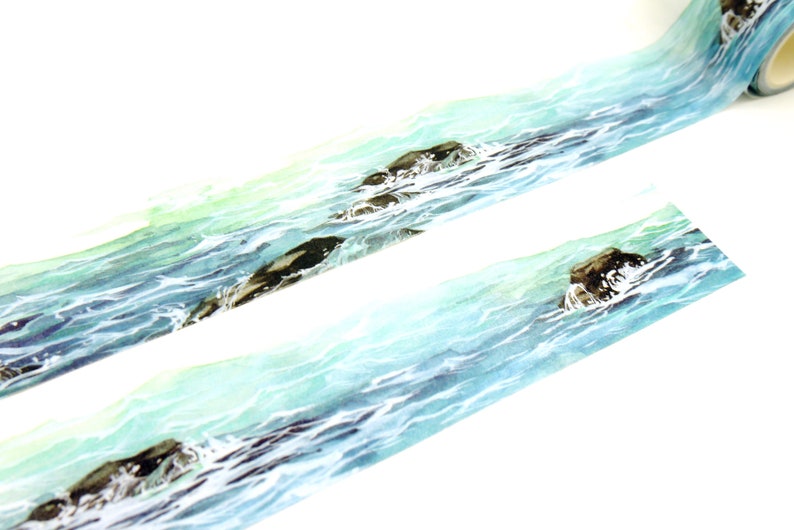 May include: A roll of washi tape with a watercolor design of ocean waves and rocks. The tape is light blue and green with brown rocks.
