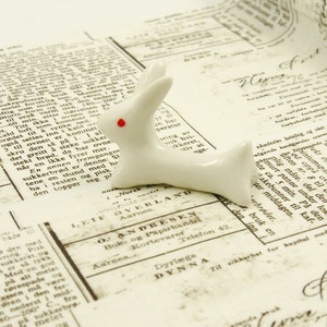 Puede incluir: Una figurita de conejo de cerámica blanca con un punto rojo en la nariz, tumbada sobre un periódico antiguo con texto en un idioma extranjero.