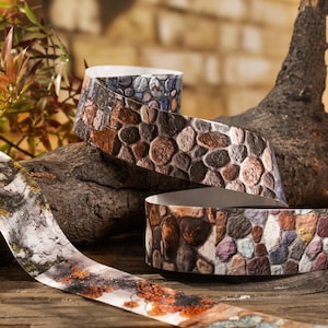 Peut inclure: Ruban d&eacute;coratif &agrave; motif de pierre dans les tons de marron, gris et bleu. Le ruban est enroul&eacute; et pr&eacute;sent&eacute; sur une surface en bois, avec un fond naturel de bois et de feuillage. Le motif du ruban imite l'aspect d'un chemin pav&eacute;.