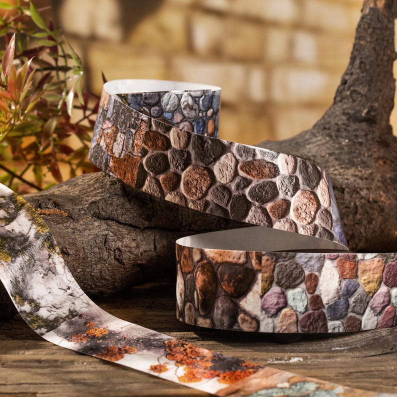 Peut inclure: Ruban d&eacute;coratif &agrave; motif de pierre dans les tons de marron, gris et bleu. Le ruban est enroul&eacute; et pr&eacute;sent&eacute; sur une surface en bois, avec un fond naturel de bois et de feuillage. Le motif du ruban imite l'aspect d'un chemin pav&eacute;.