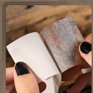Peut inclure: Gros plan sur des mains tenant un petit morceau de papier rectangulaire. Le papier est en train d'&ecirc;tre d&eacute;coll&eacute; pour r&eacute;v&eacute;ler une surface &agrave; motifs avec des nuances de rouge, de marron et de gris. Les ongles de la personne sont peints en noir.