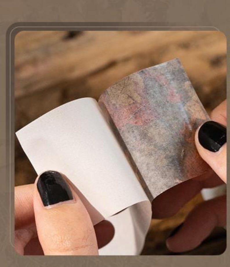 Peut inclure: Gros plan sur des mains tenant un petit morceau de papier rectangulaire. Le papier est en train d'&ecirc;tre d&eacute;coll&eacute; pour r&eacute;v&eacute;ler une surface &agrave; motifs avec des nuances de rouge, de marron et de gris. Les ongles de la personne sont peints en noir.