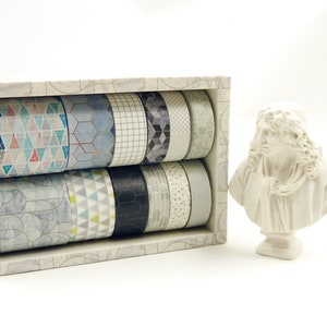 Pode incluir: Um conjunto de 12 fitas washi numa caixa branca. As fitas têm padrões geométricos em tons de azul, cinzento e branco. A caixa tem um padrão dourado e branco.
