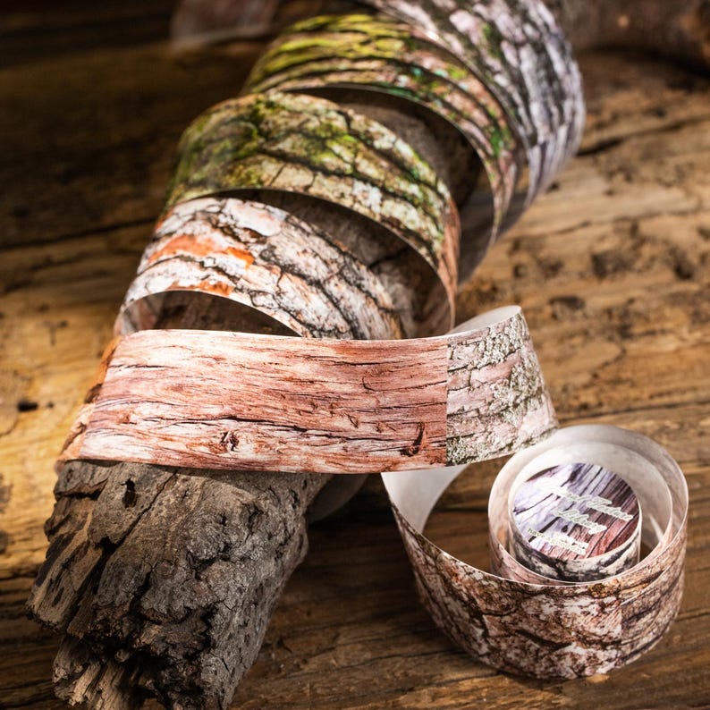 Peut inclure: Un rouleau de papier d&eacute;coratif avec une texture d'&eacute;corce dans diverses couleurs, dont le marron, le rouge et le vert. Le papier est enroul&eacute; et dispos&eacute; sur une surface en bois, mettant en valeur les motifs d&eacute;taill&eacute;s du grain du bois. Le papier mesure environ 5 cm de large.