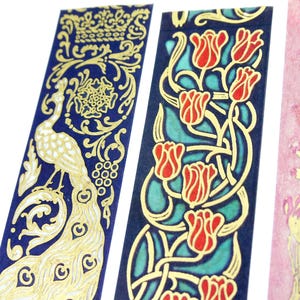 Pu&ograve; includere: Tre rotoli di nastro washi decorativo con disegni in foglia d'oro. Il primo rotolo presenta un pavone su sfondo blu. Il secondo rotolo ha un disegno floreale con tulipani rossi e uno sfondo blu e verde. Il terzo rotolo ha uno sfondo rosa con fiori in foglia d'oro.