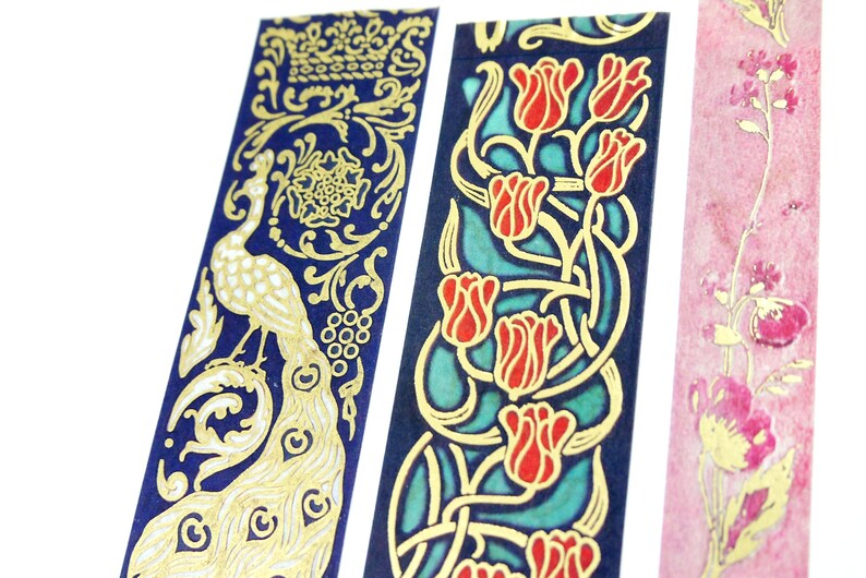 Pu&ograve; includere: Tre rotoli di nastro washi decorativo con disegni in foglia d'oro. Il primo rotolo presenta un pavone su sfondo blu. Il secondo rotolo ha un disegno floreale con tulipani rossi e uno sfondo blu e verde. Il terzo rotolo ha uno sfondo rosa con fiori in foglia d'oro.