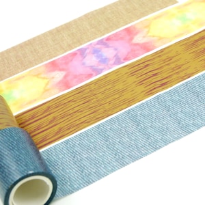 Op de afbeelding: Vier rollen plakband gerangschikt in een verticale stapel. De patronen omvatten een beige linnen textuur, een levendige aquarelwas in roze, geel en blauw, een bruin houtnerfpatroon en een donkerblauwe spijkerstof textuur.