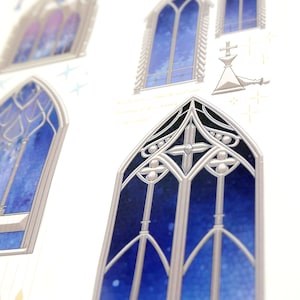 May include: Close-up of a sheet of stickers featuring illustrations of gothic cathedral windows. The windows have a blue and purple stained-glass effect, with silver-coloured frames. Some windows have a cross design.