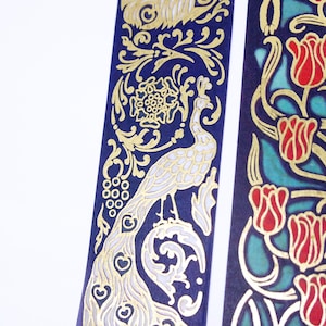 Pu&ograve; includere: Due rotoli di nastro washi decorativo con disegni dorati e bianchi su sfondo blu scuro. Un rotolo presenta un disegno di pavone e l'altro un disegno floreale con tulipani rossi.