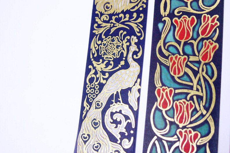 Pu&ograve; includere: Due rotoli di nastro washi decorativo con disegni dorati e bianchi su sfondo blu scuro. Un rotolo presenta un disegno di pavone e l'altro un disegno floreale con tulipani rossi.