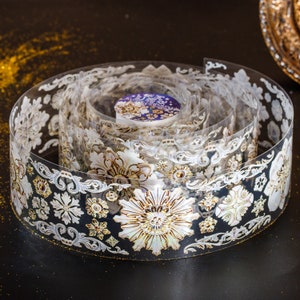 May include: A roll of decorative washi tape with a white and gold floral design. The tape is rolled up and the design is visible on the outside of the roll.