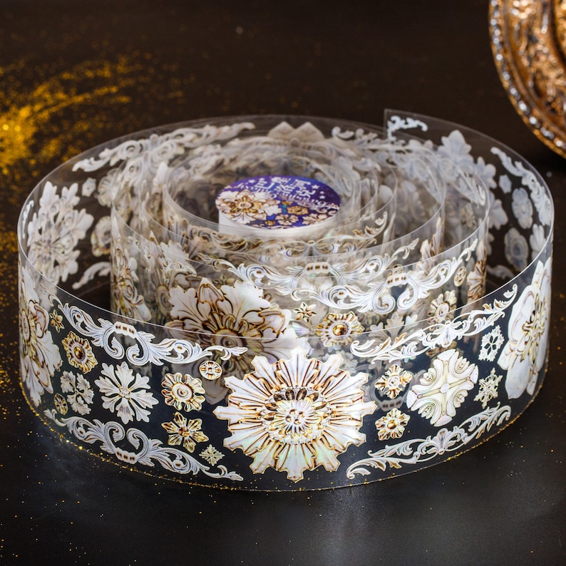 May include: A roll of decorative washi tape with a white and gold floral design. The tape is rolled up and the design is visible on the outside of the roll.