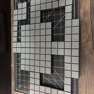 Op de afbeelding: Opstelling van grijze tegels met baksteenpatroon en zwarte randen, gemonteerd op een zwarte snijmat met witte rasterlijnen en inch-maten. De tegels zijn gerangschikt in een rechthoekige vorm met een uitsparing.