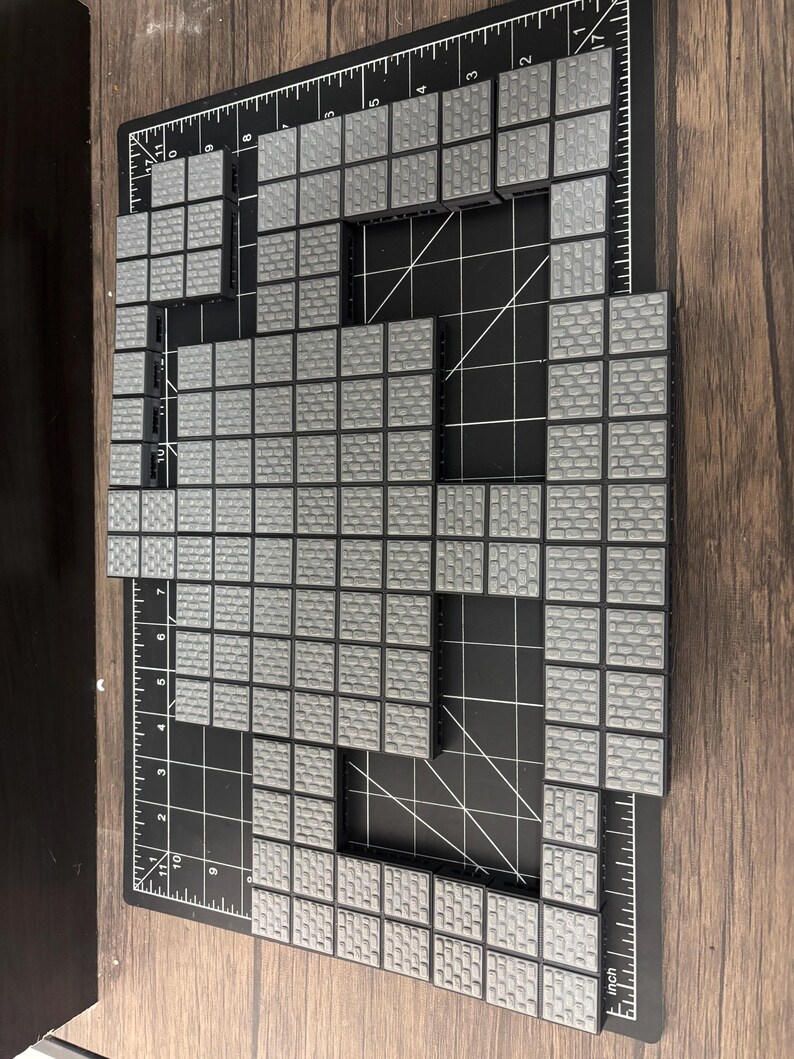 Op de afbeelding: Opstelling van grijze tegels met baksteenpatroon en zwarte randen, gemonteerd op een zwarte snijmat met witte rasterlijnen en inch-maten. De tegels zijn gerangschikt in een rechthoekige vorm met een uitsparing.