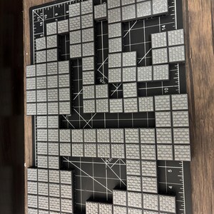 Op de afbeelding: Een verzameling grijze, vierkante tegels gerangschikt op een zwarte snijmat. De tegels zijn ontworpen om op metselwerk te lijken. De opstelling suggereert een gepixelde afbeelding of een bouwsteenconstructieproject. De mat heeft inch-markeringen.