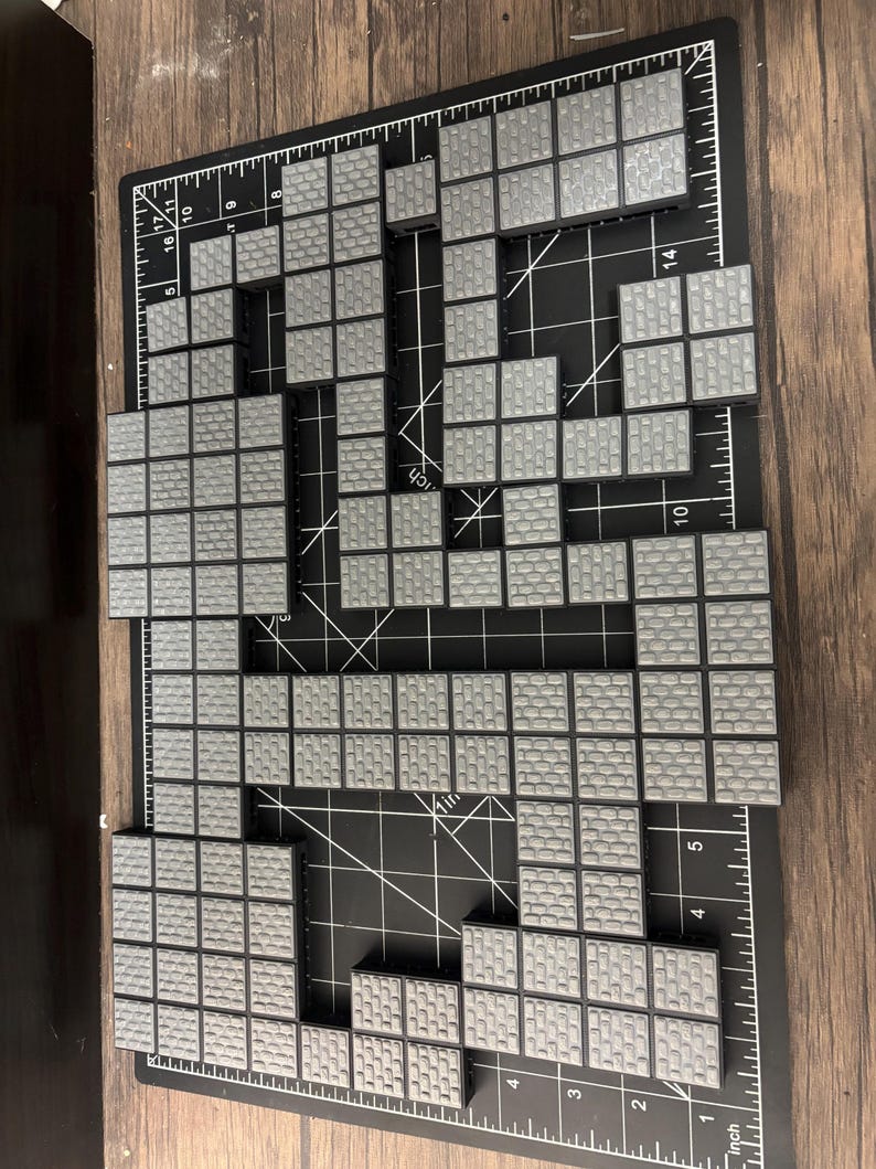Op de afbeelding: Een verzameling grijze, vierkante tegels gerangschikt op een zwarte snijmat. De tegels zijn ontworpen om op metselwerk te lijken. De opstelling suggereert een gepixelde afbeelding of een bouwsteenconstructieproject. De mat heeft inch-markeringen.