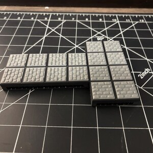 Op de afbeelding: Grijze plastic terreintegels met verschillende steenstructuren. De tegels zijn in een L-vorm gerangschikt, met een zwarte basis. Ontworpen voor tabletop gaming en miniature wargaming.