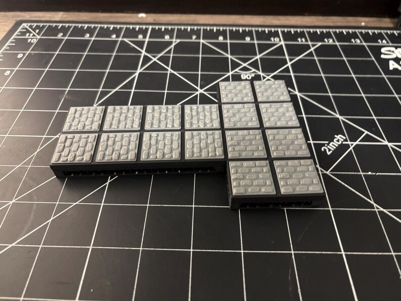 Op de afbeelding: Grijze plastic terreintegels met verschillende steenstructuren. De tegels zijn in een L-vorm gerangschikt, met een zwarte basis. Ontworpen voor tabletop gaming en miniature wargaming.