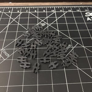 Op de afbeelding: Een verzameling kleine, zwarte plastic connectoren verspreid op een zwarte snijmat met witte rasterlijnen en afmetingen. De connectoren hebben een unieke vorm met twee afgeronde uiteinden.