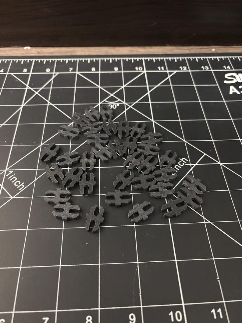 Op de afbeelding: Een verzameling kleine, zwarte plastic connectoren verspreid op een zwarte snijmat met witte rasterlijnen en afmetingen. De connectoren hebben een unieke vorm met twee afgeronde uiteinden.