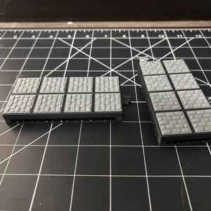 Op de afbeelding: Twee grijze, rechthoekige miniatuur terreinstukken met een kasseien textuur. De stukken zijn gemaakt van een donkergrijs materiaal en zijn ontworpen voor tabletop gaming of modelbouw. De stukken liggen op een zwarte snijmat.