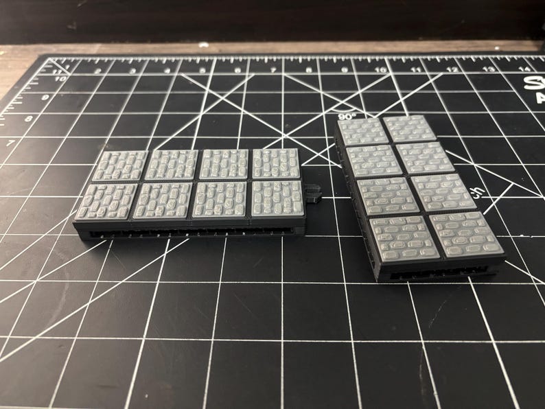 Op de afbeelding: Twee grijze, rechthoekige miniatuur terreinstukken met een kasseien textuur. De stukken zijn gemaakt van een donkergrijs materiaal en zijn ontworpen voor tabletop gaming of modelbouw. De stukken liggen op een zwarte snijmat.